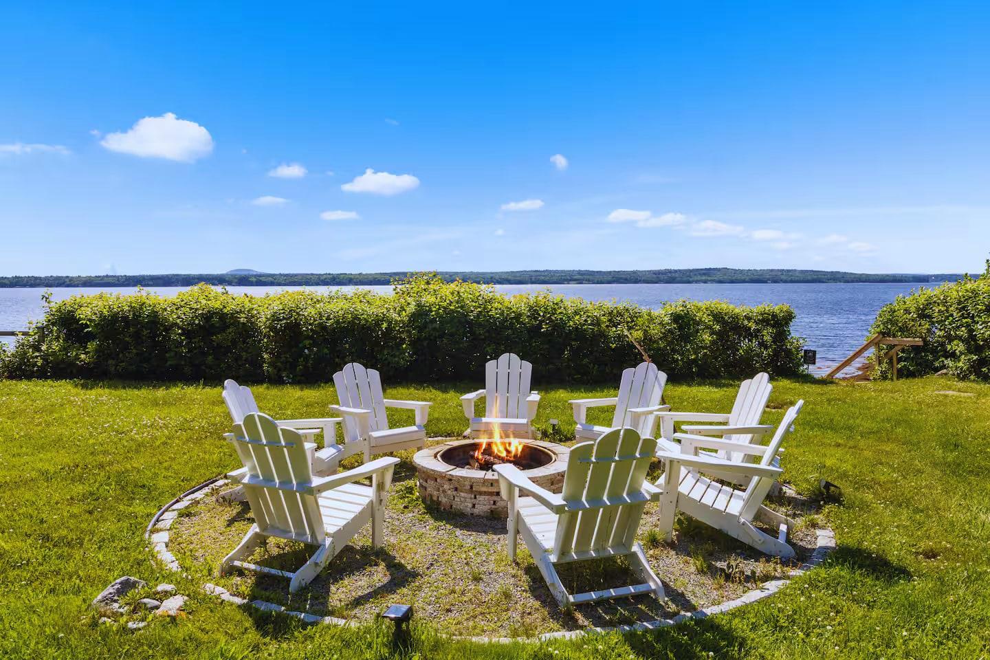Fire pit circle with Adirondack chairs on a summer day, the bay beyond the hedge at Eagles Nest